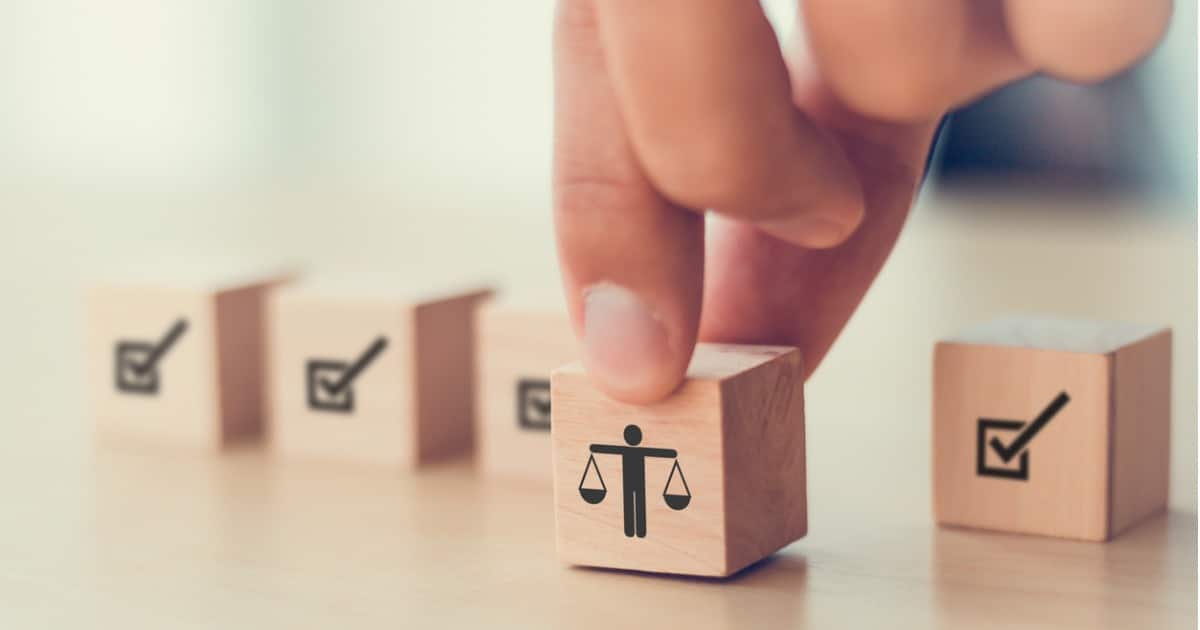 A hand holding a wooden block. The block has an icon of a man holding a balance scale. The background has four more blocks with checkbox icons.