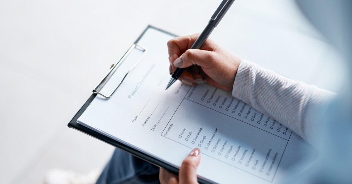 Close up of woman's hand holding a pen and filling out a form that appears to be a medical history form. The form is titled Patient Questionnaire and there are spaces to fill out name, DOB and a list of symptoms to check.